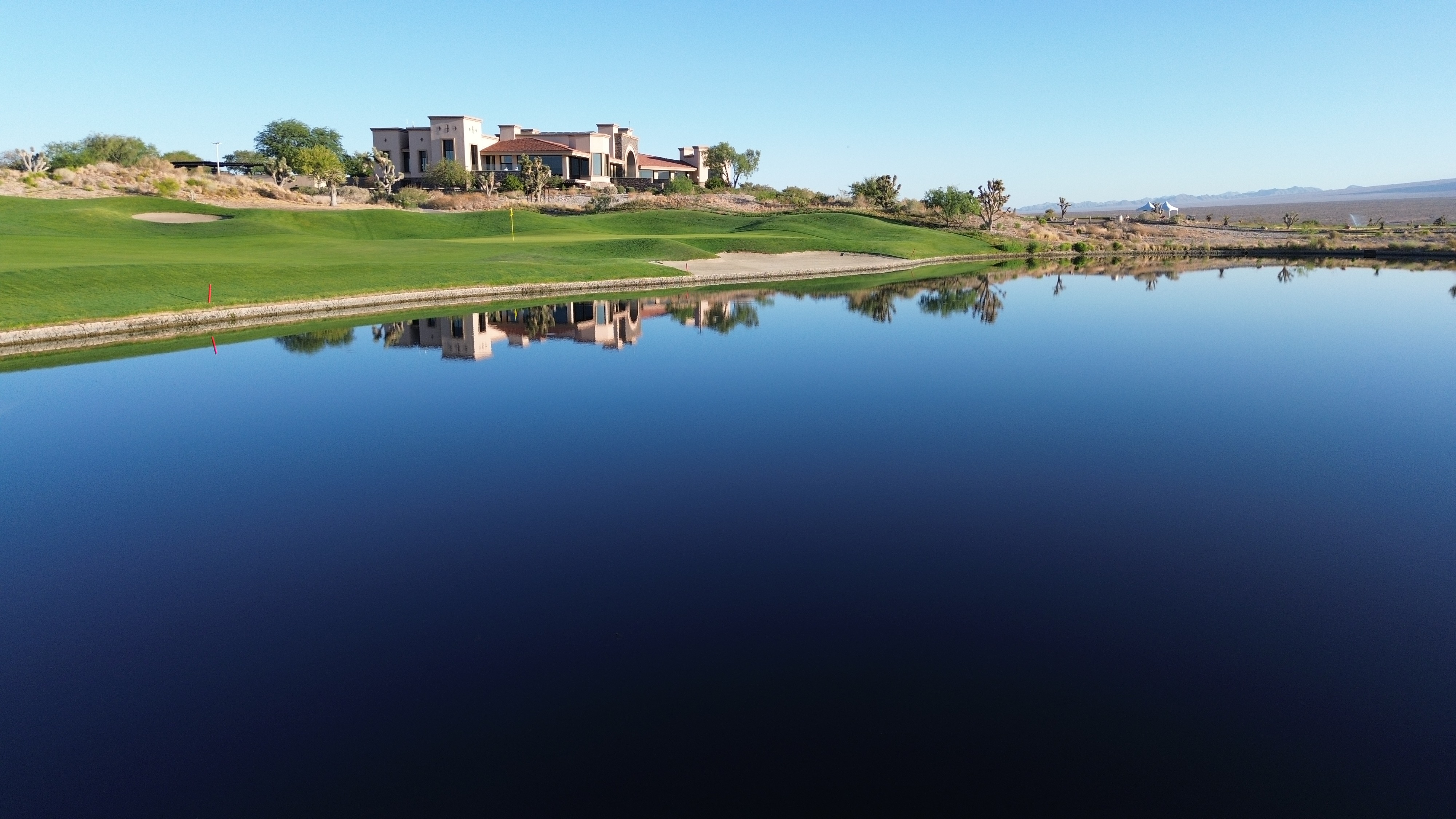 Desert golf course clubhouse reflected in a calm lake with a manicured green and distant mountains